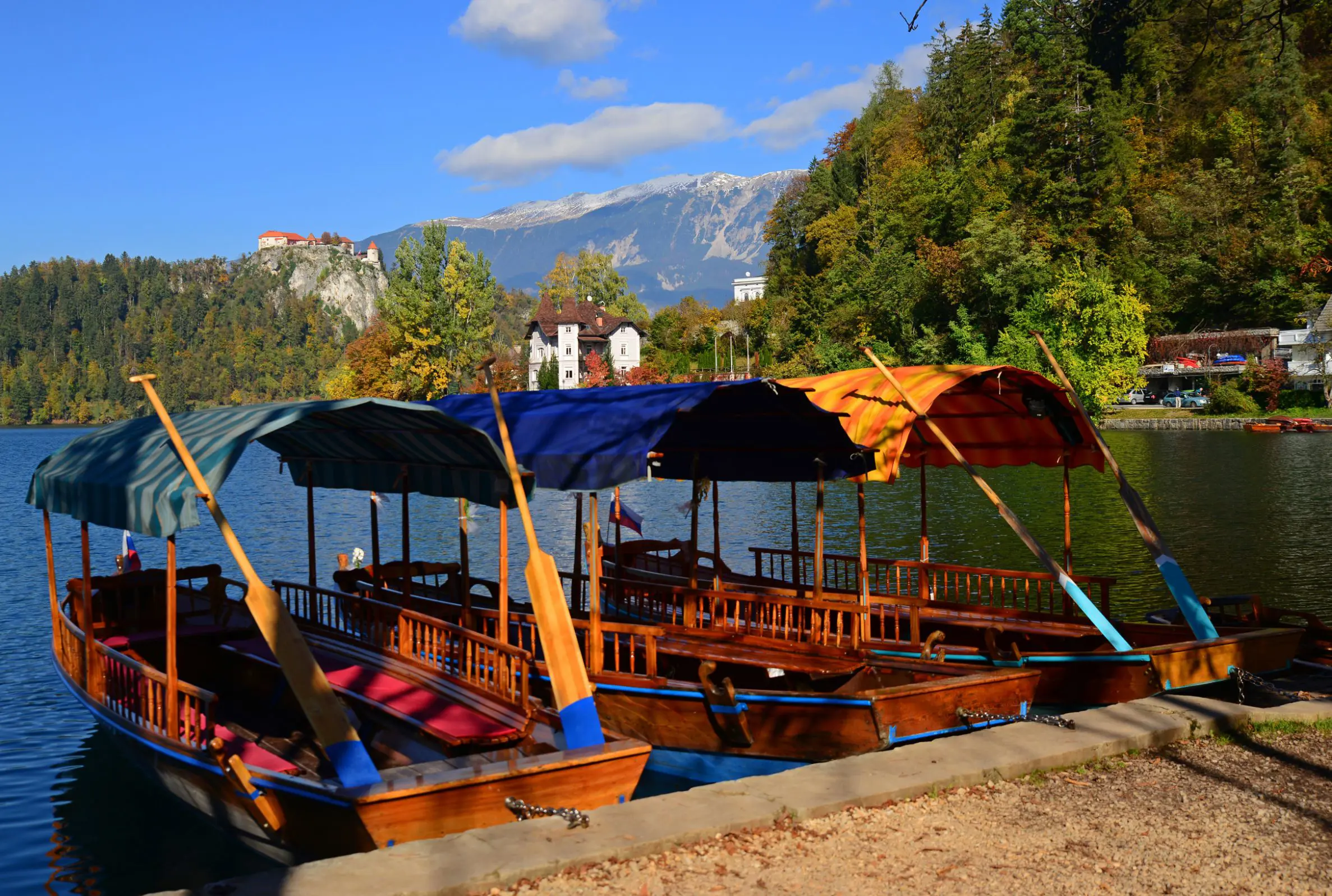 Back of two pletna boats docked on Lake Bled, with forests on the other side. A large building on a hill and one on ground level, and mountains in the distance.