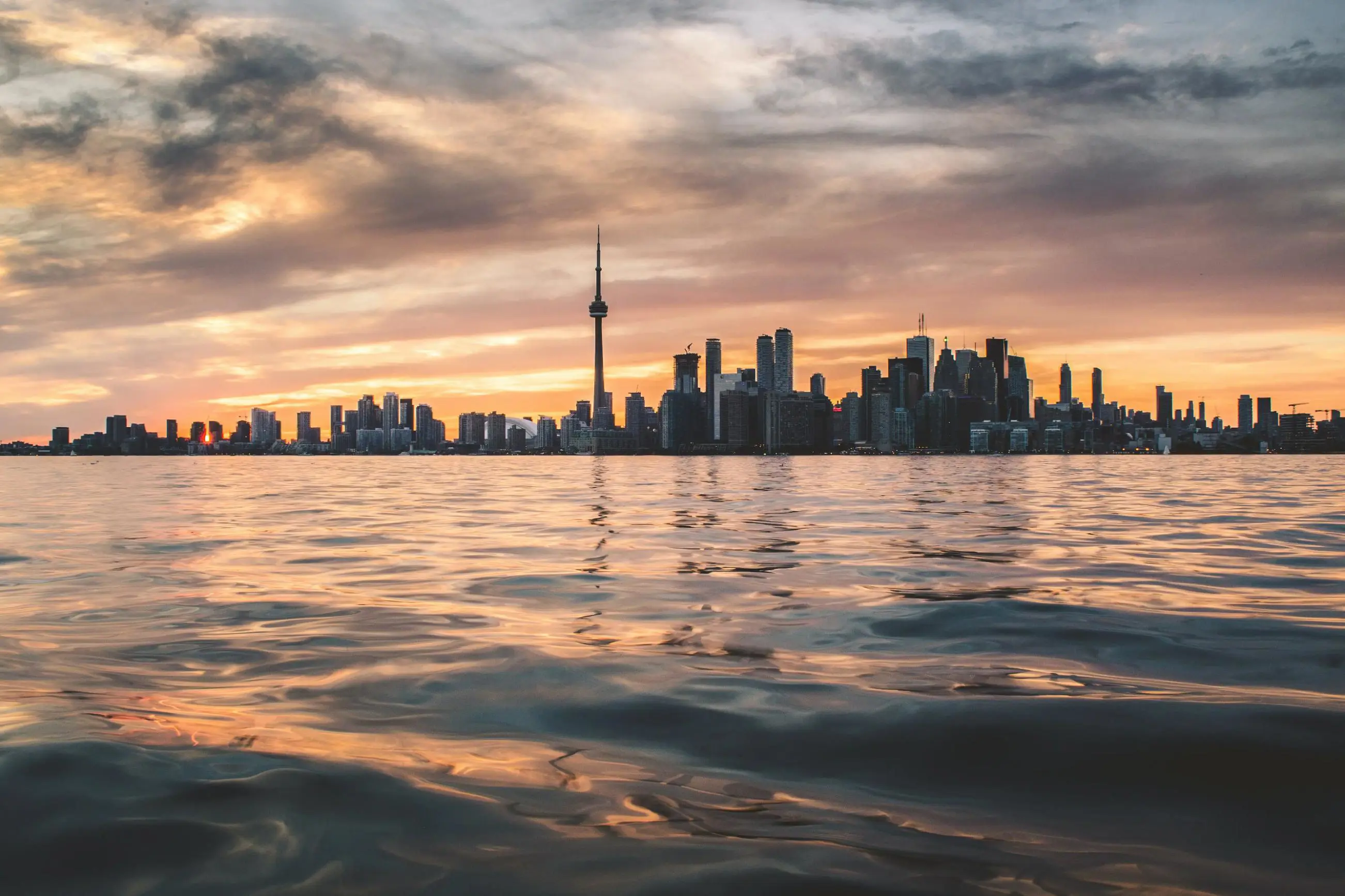 View over the water towards the Toronto skyline at sunset, with the CN Tower and surrounding buildings silhouetted against a pink and orange sky