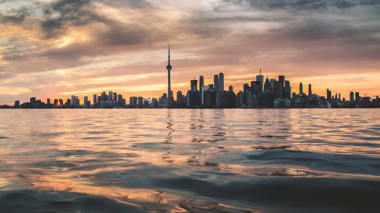 View over the water towards the Toronto skyline at sunset, with the CN Tower and surrounding buildings silhouetted against a pink and orange sky