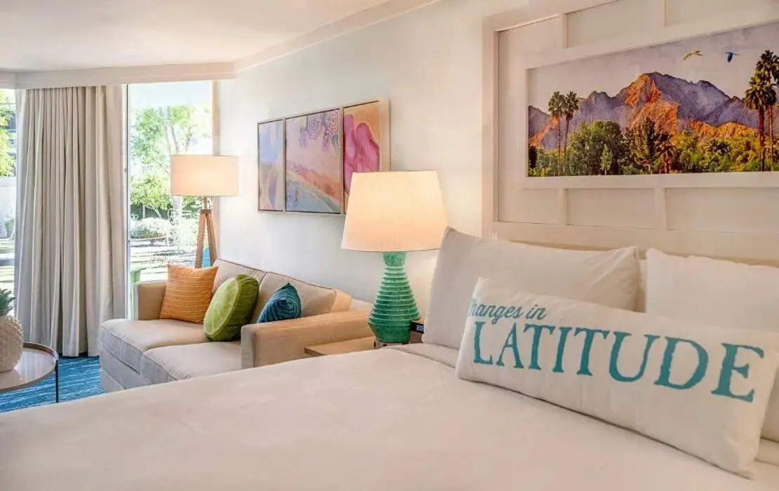 Stylish guest bedroom at the Riviera Resort & Spa, Palm Springs, featuring a large bed with crisp white linens, colourful décor, a seating area, and floor-to-ceiling windows with views of the surrounding landscape
