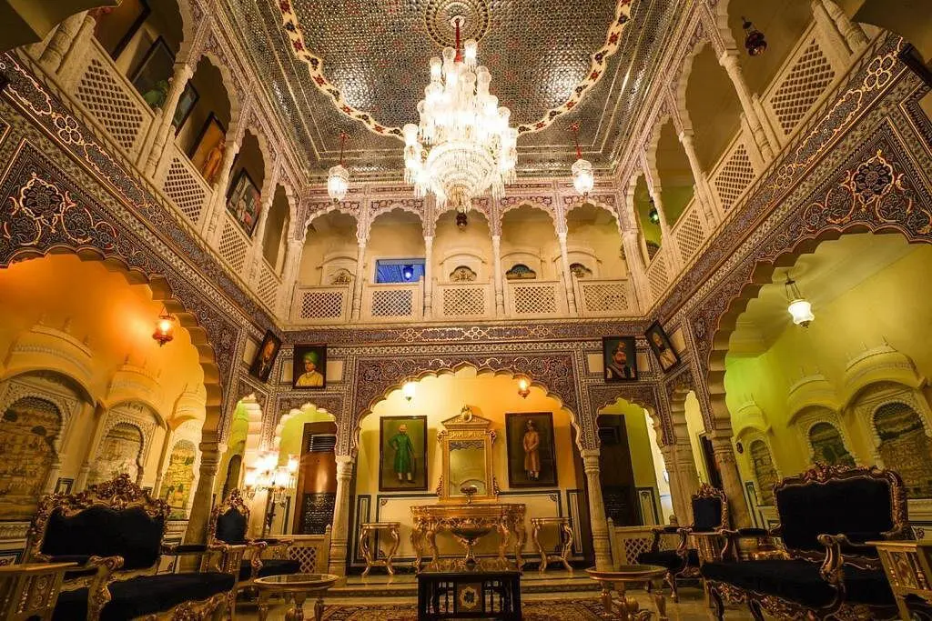 The ornate interior of Shahpura House, Jaipur, with intricately carved arches, a mosaic ceiling, a grand crystal chandelier, and royal portraits