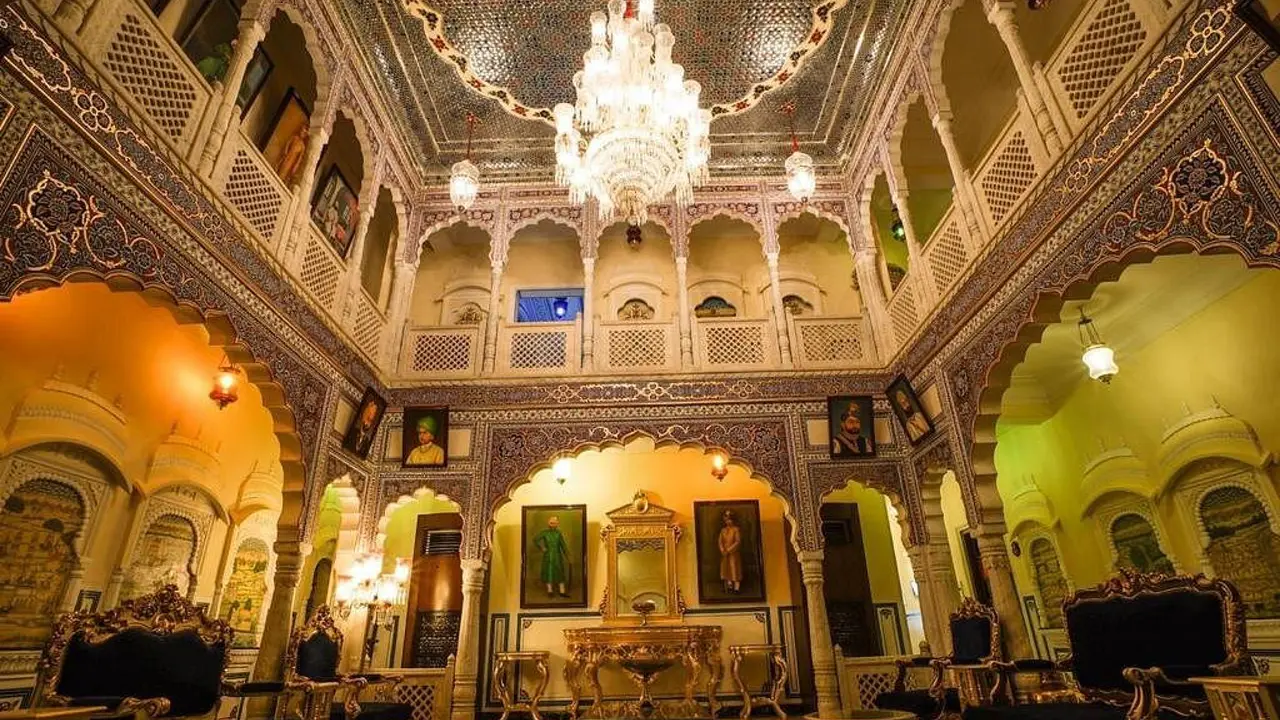 The ornate interior of Shahpura House, Jaipur, with intricately carved arches, a mosaic ceiling, a grand crystal chandelier, and royal portraits