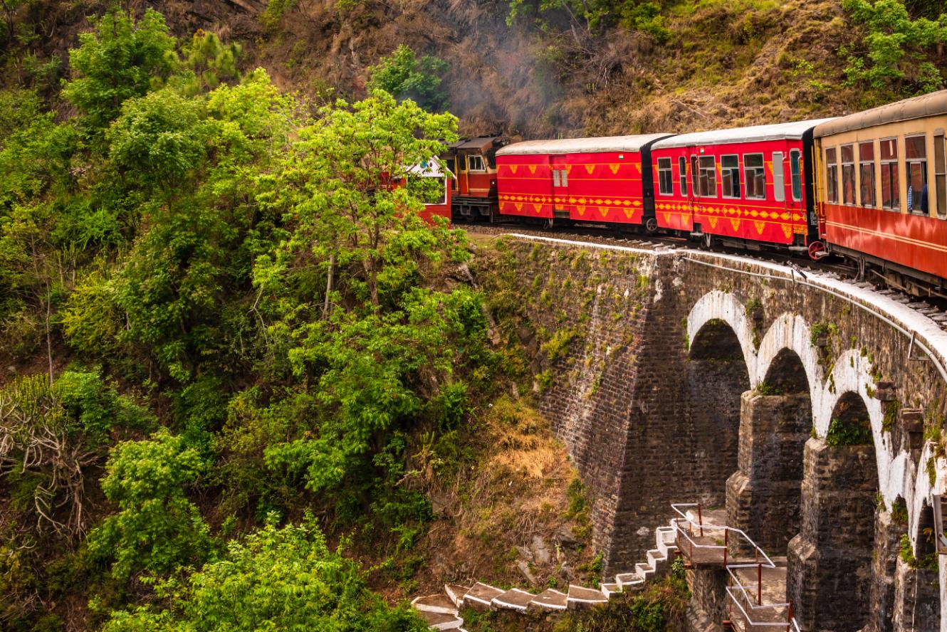 The heritage toy train crossing a railway bridge on the Kalka–Shimla route, surrounded by lush green hills and trees