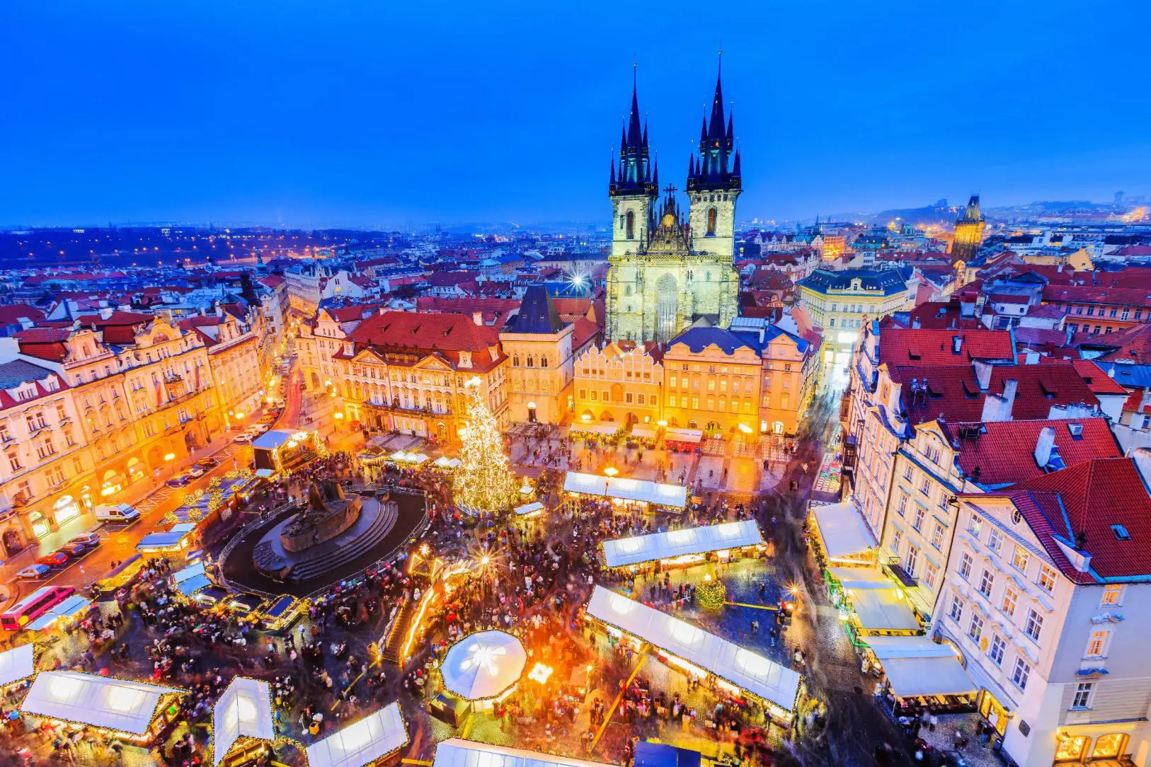 High angle shot of a Christmas market in the town square of Prague, showing a lit up gothic church with two towers and a large lit up Christmas tree