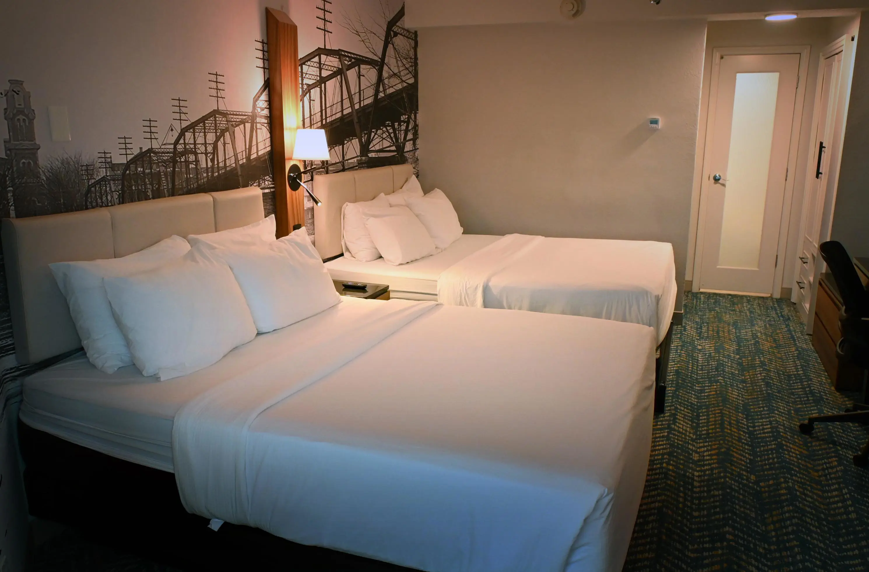 Double room at Fort Pontchartrain hotel, featuring two large beds with white linens, a monochrome mural of a historic bridge, bedside lighting, and a modern interior