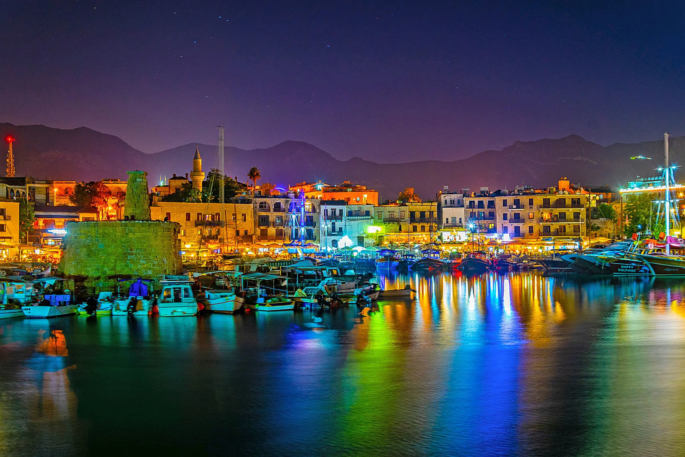 View of a harbour at night, with boats docked and a ruin to the left. Multi coloured lights behind the boats, reflecting on the water. A strip of buildings on the far side of the harbour, in front of a silhouette of mountains in front of a night sky.