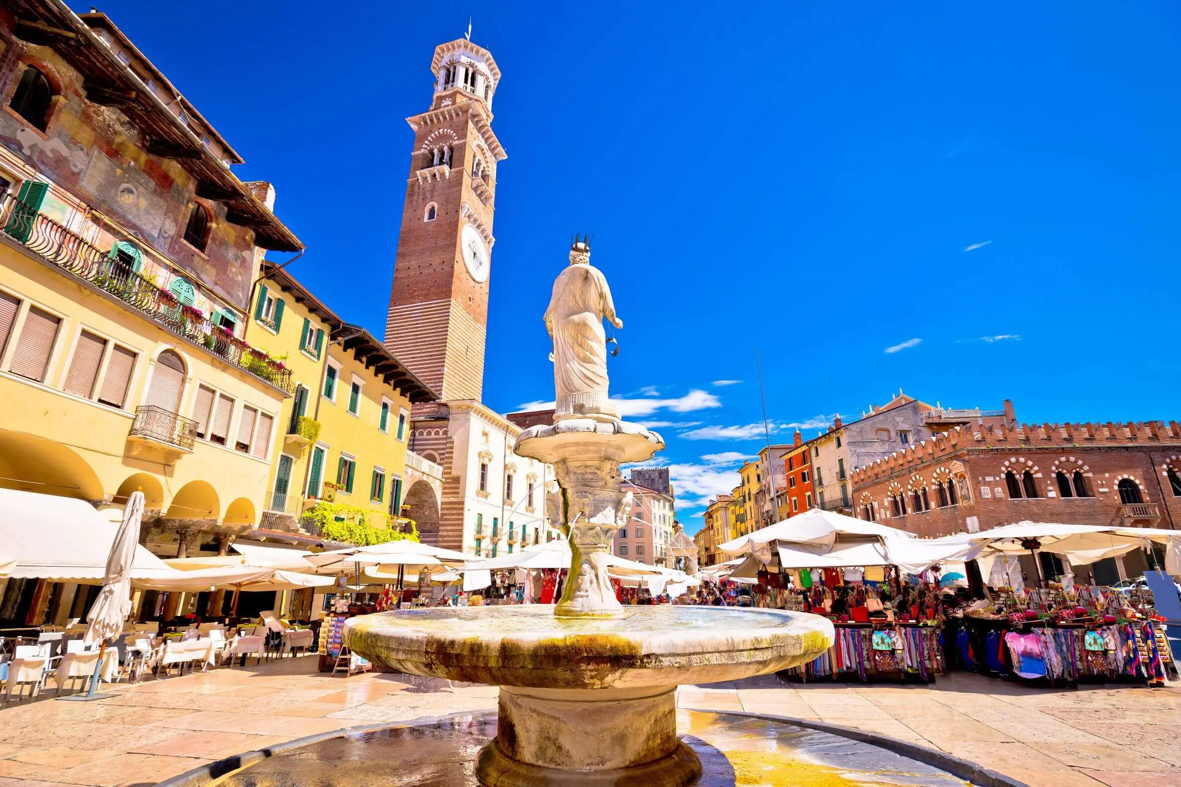 View of Fontana Madonna fountain from behind, showing the Torre dei Lamberti tower and the square's restaurants and markets