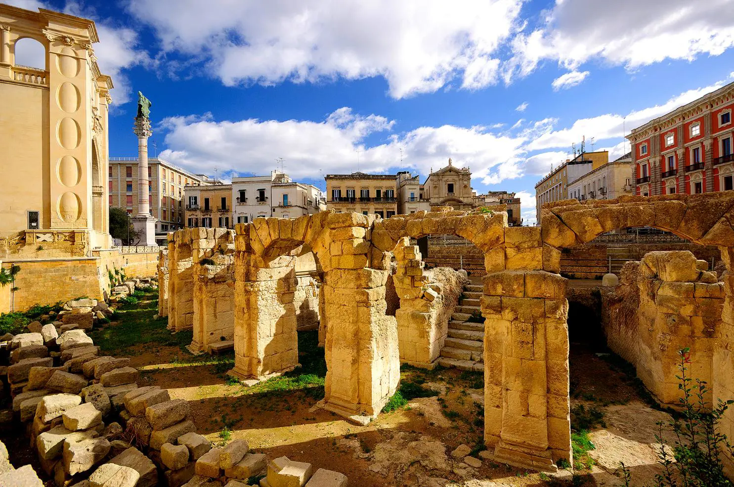 Ancient Roman ruins in the city centre of Lecce, Puglia, Italy, with weathered stone arches and columns surrounded by historic buildings under a bright blue sky with scattered clouds