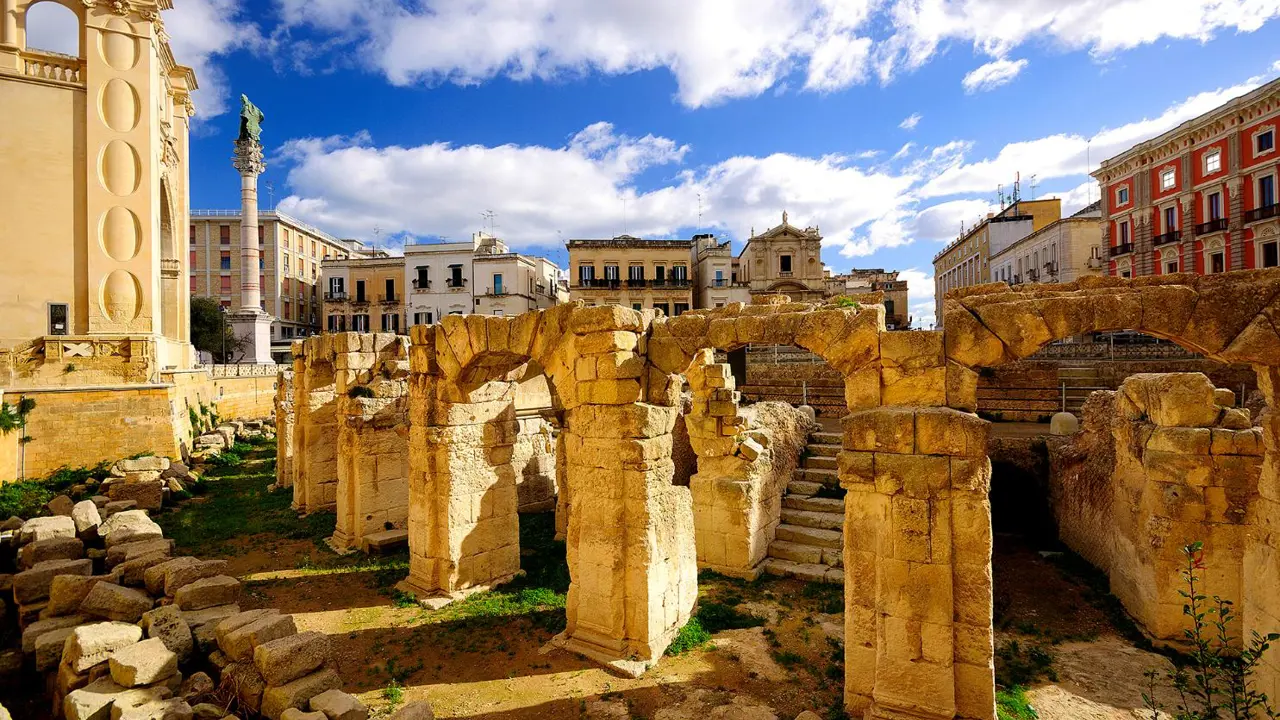 Ancient Roman ruins in the city centre of Lecce, Puglia, Italy, with weathered stone arches and columns surrounded by historic buildings under a bright blue sky with scattered clouds
