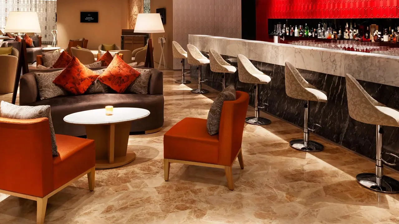 The bar area at the Leela Ambience Convention Hotel, Delhi, with red, white, and wooden décor, a modern bar counter, comfortable seating, and warm ambient lighting