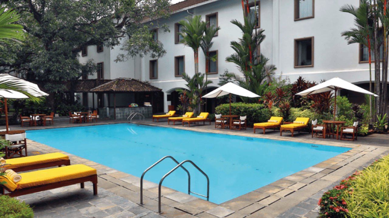 Outdoor pool at The Fern hotel in Kochi, with yellow sunbeds and white parasols, set against a white building and framed by tall palm trees