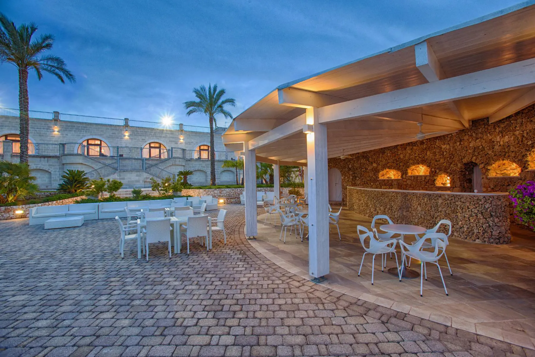 Outdoor dining and bar area at Masseria Caselli Hotel, Italy, with stone walls, white furniture, and warm evening lighting