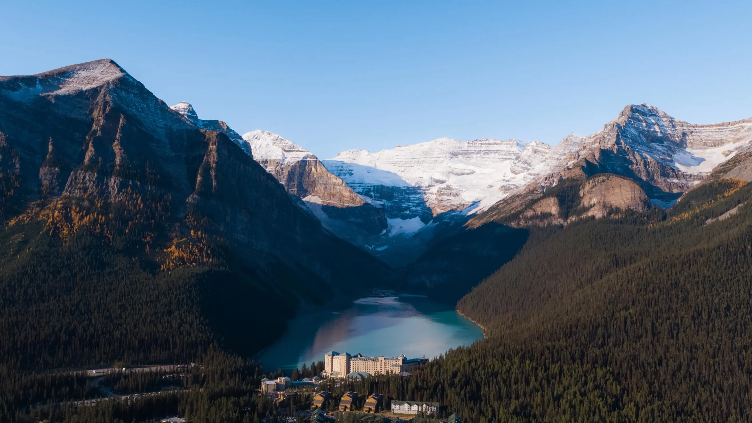 Aerial view of Fairmont Chateau, Lake Louise nestled beside a turquoise lake, surrounded by dense pine forests and towering snow-capped mountains under a clear blue sky