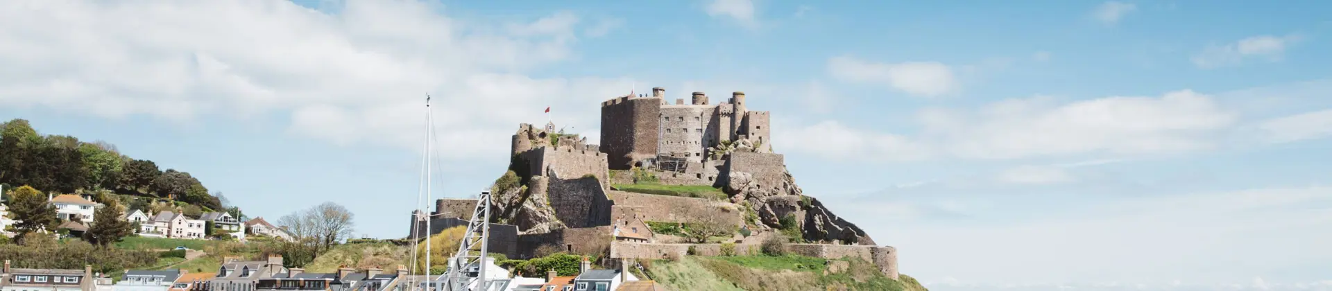 Long shot of Mont Orgueil Castle, showing part of the harbour below it and two people sat on the wall between the harbour and the sea, the buildings beside the harbour and the houses on a hill behind it, on a sunny day with a blue sky 