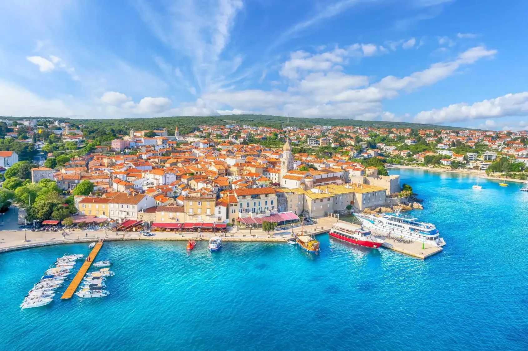 Aerial view of Krk town on the Island of Krk, Croatia, featuring orange-roofed buildings, a harbour with boats, and clear blue sea