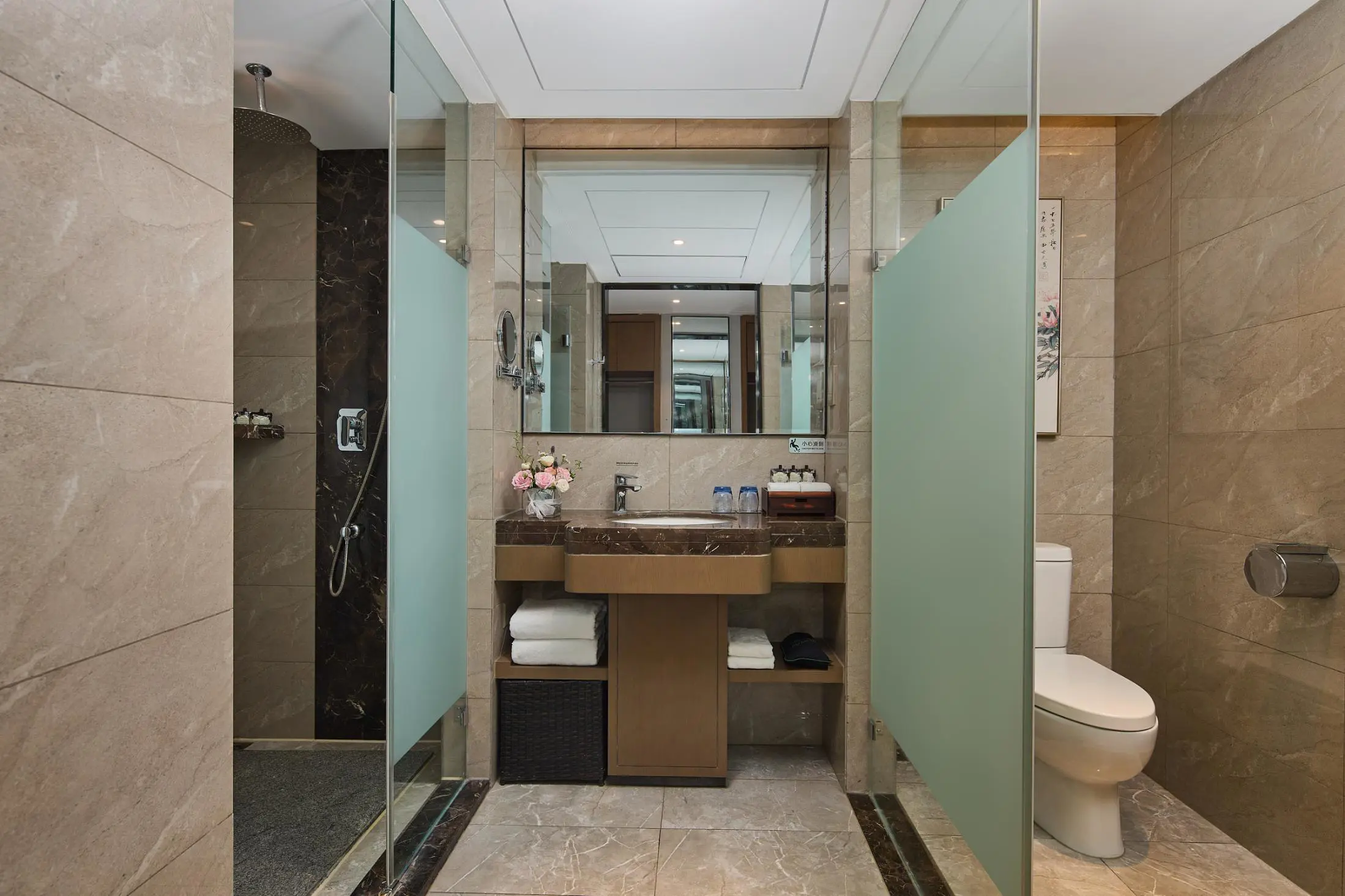Modern bathroom at Youngor Central Hotel with marble-style tiles, a glass-enclosed shower, a vanity with a large mirror, neatly arranged amenities and a toilet separated by frosted glass panels