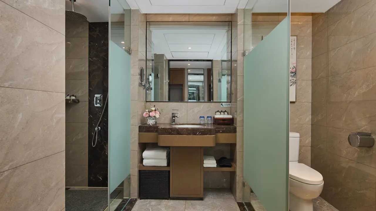 Modern bathroom at Youngor Central Hotel with marble-style tiles, a glass-enclosed shower, a vanity with a large mirror, neatly arranged amenities and a toilet separated by frosted glass panels