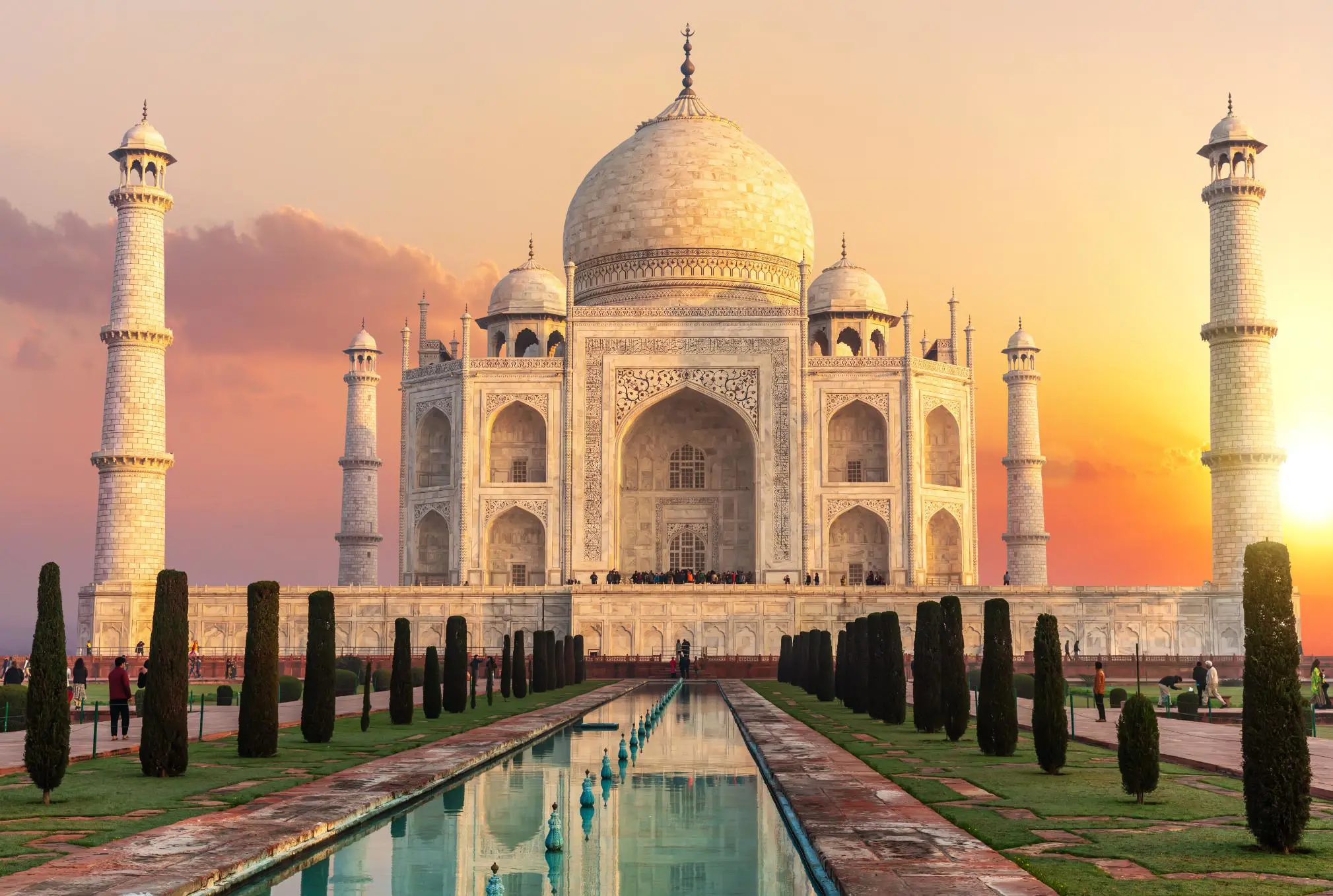 The Taj Mahal in Agra glowing in the warm hues of sunset, its white marble reflecting the golden light, with the surrounding minarets and the long reflecting pool in front adding to its majestic symmetry