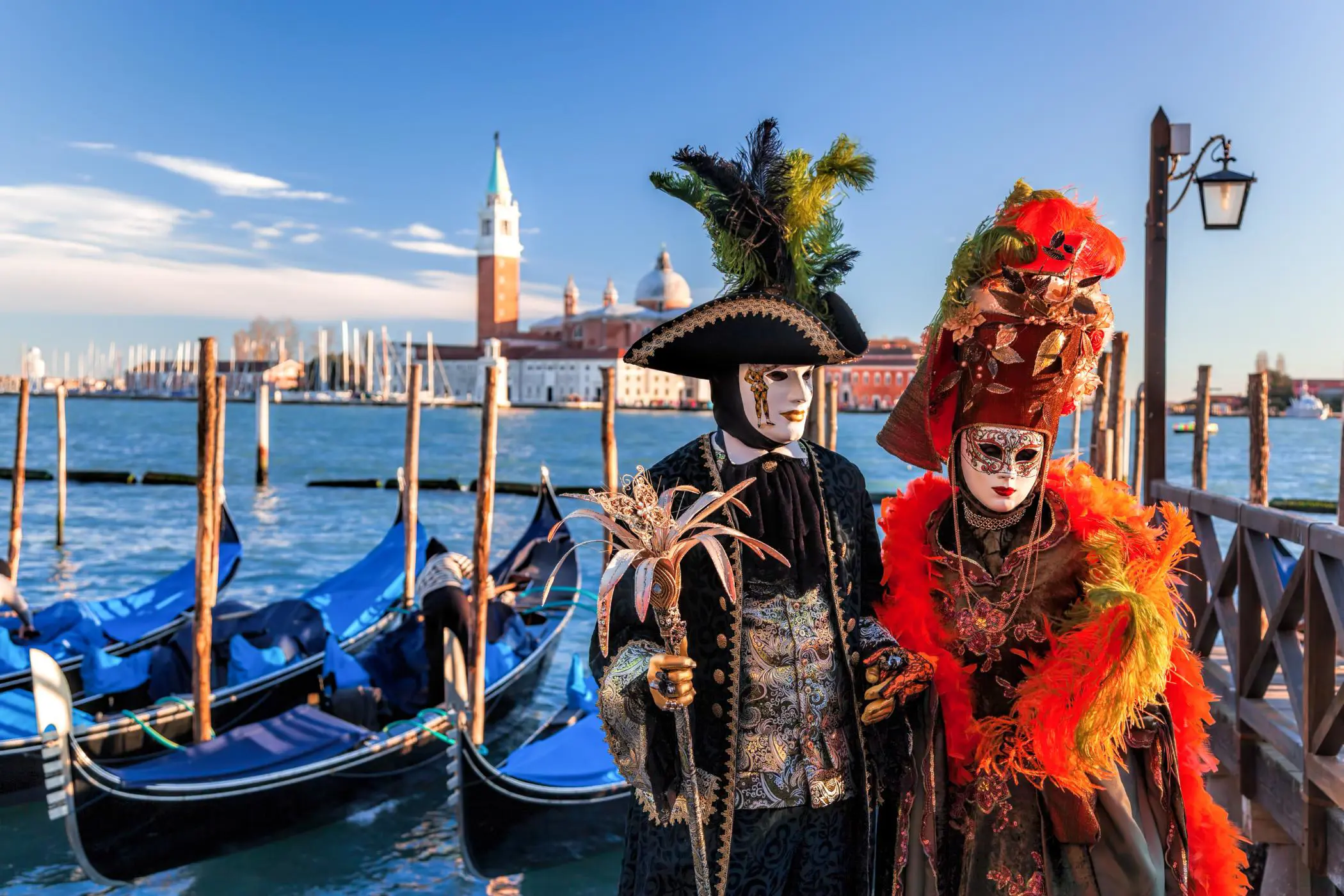 Two people in elaborate Venetian carnival costumes and masks standing by gondolas on a canal, with the Church of San Giorgio Maggiore in the background