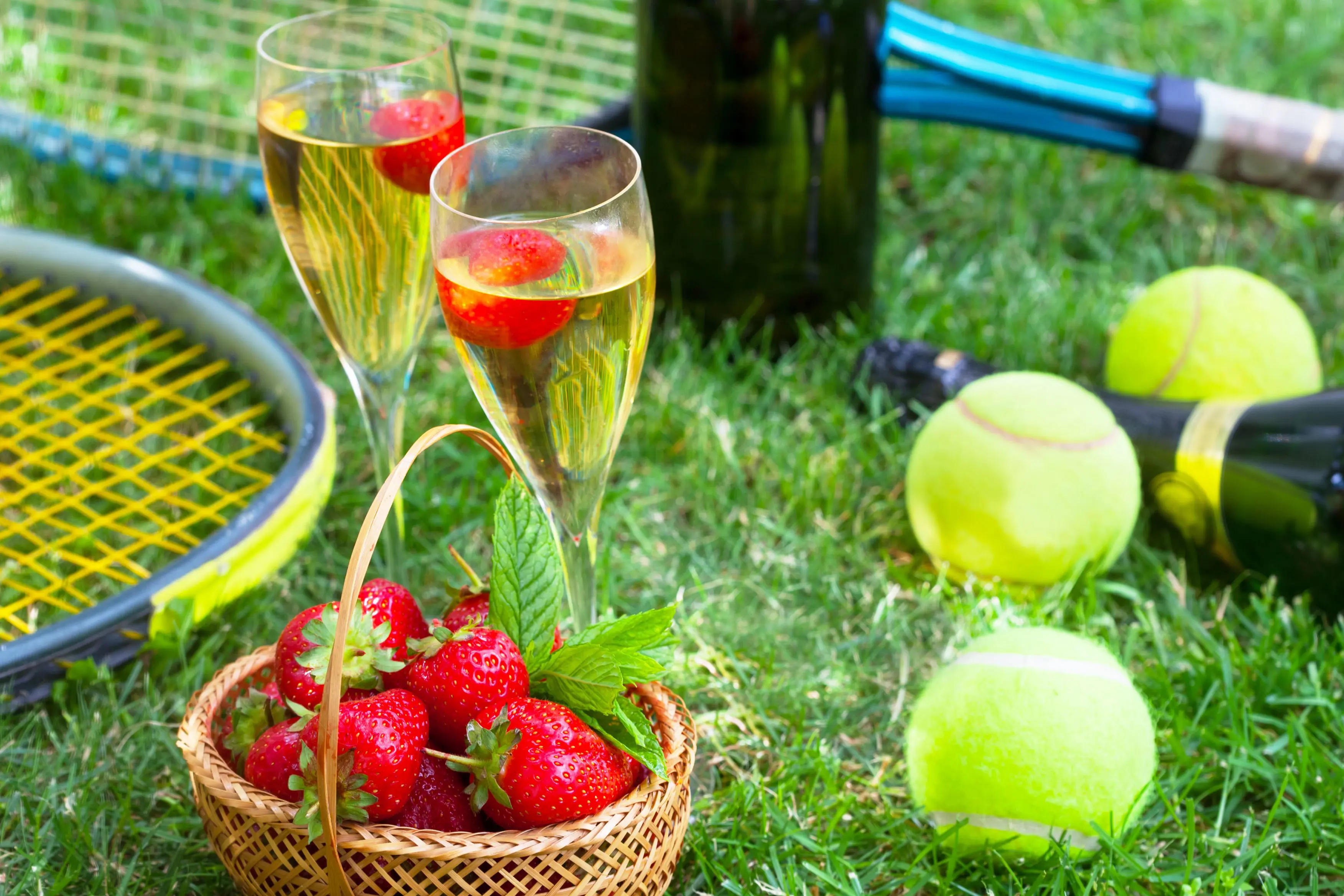 Close up of two glasses of champagne, a basket of strawberries, tennis balls and rackets on some grass