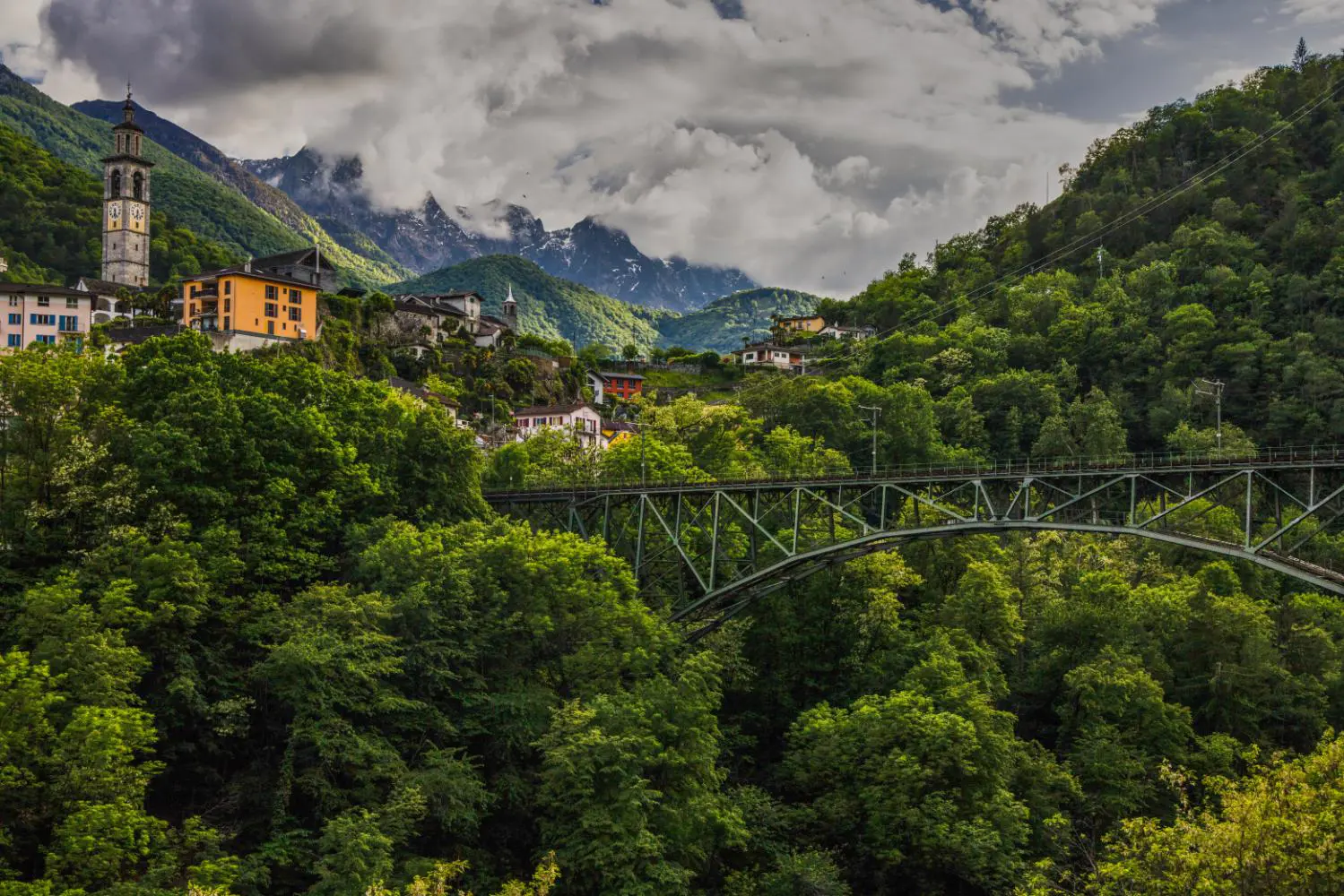 Landscape view of the Centovalli Railway bridge, surrounded by tall green trees, with mountains and a distant church tower in the background