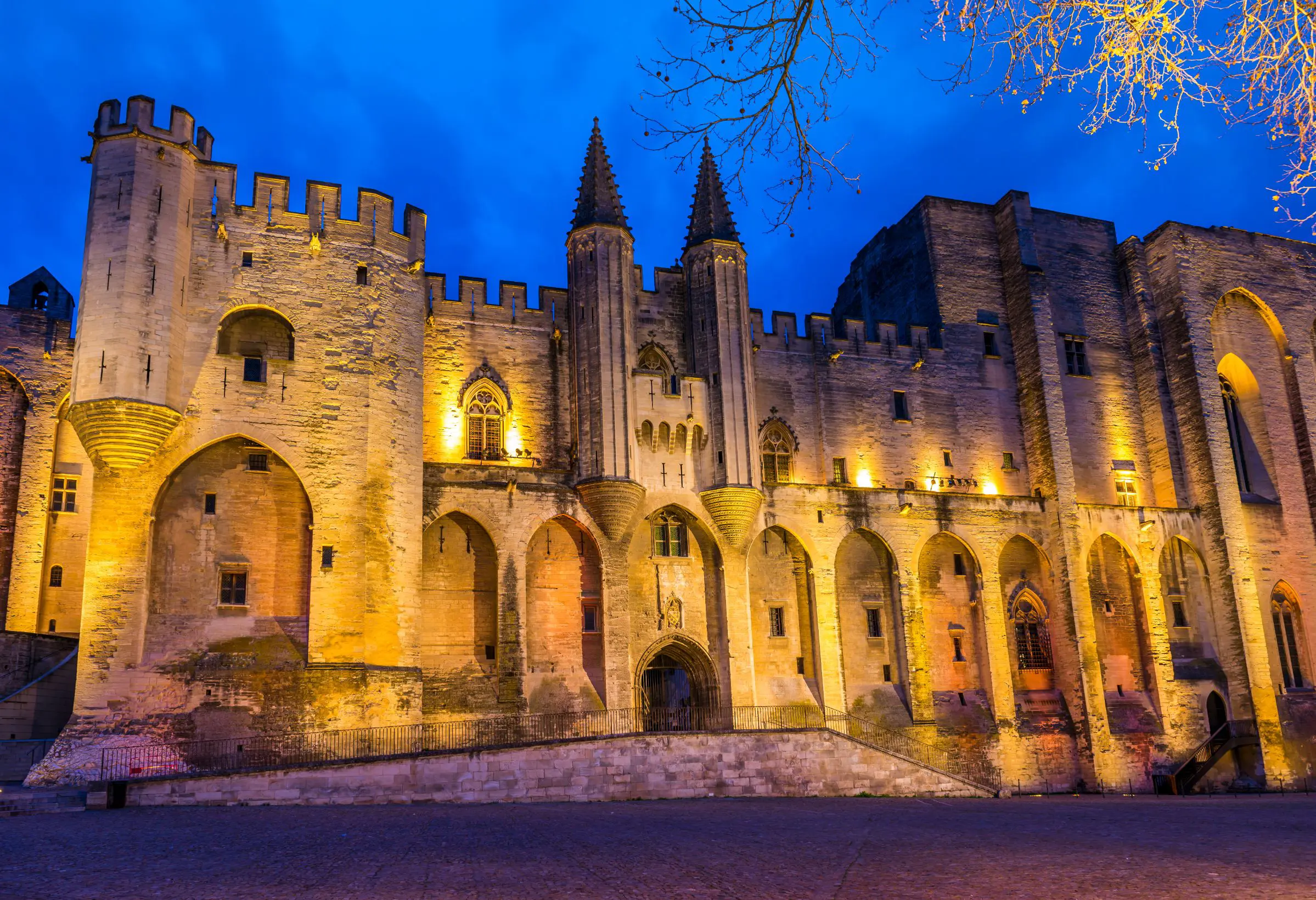 Low angle shot of a beige coloured stone palace, lit up with amber lights on the front. With pointy turrets in the middle and battlements on the left side. In front of a bright blue night sky.