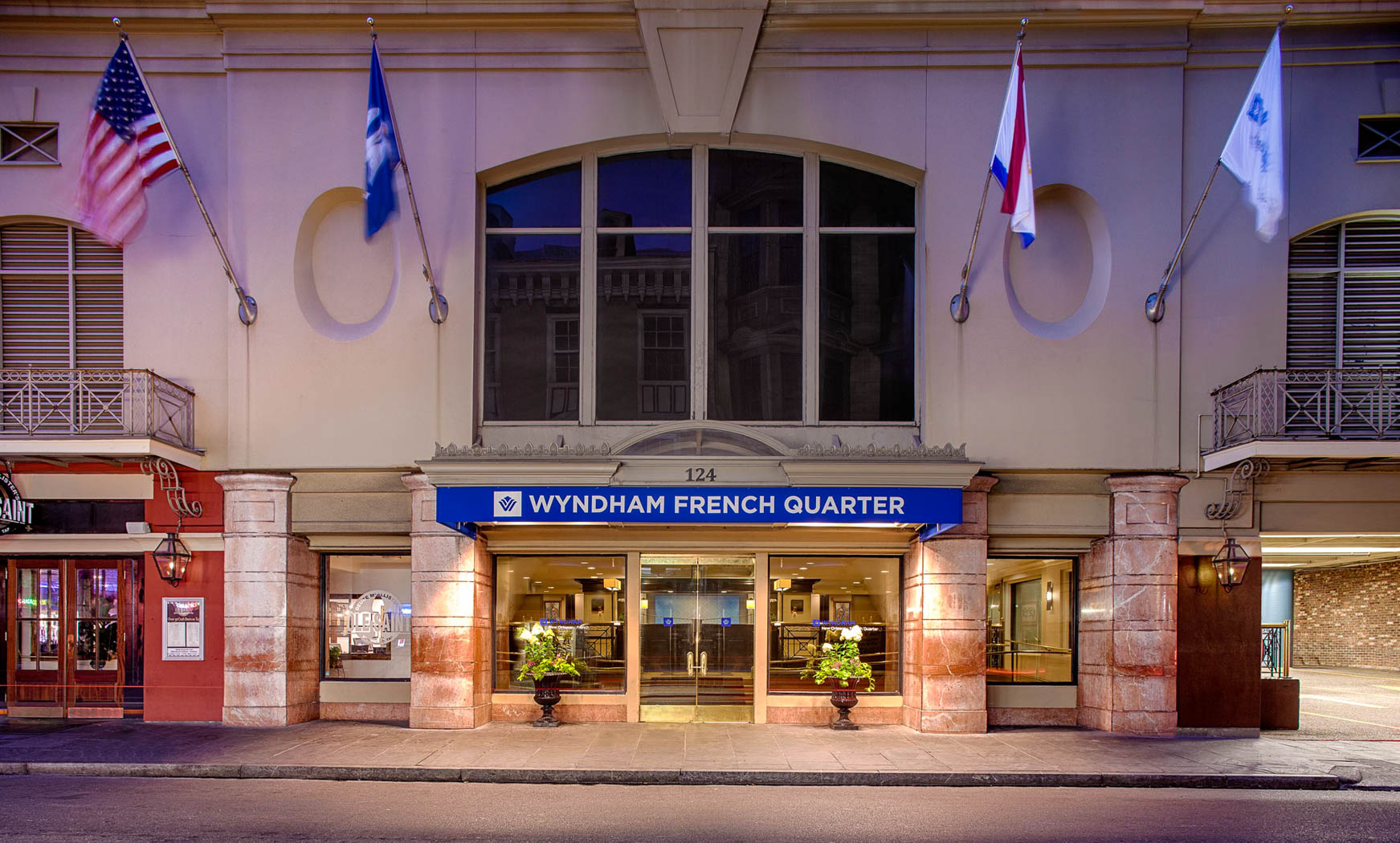 Entrance to the Wyndham French Quarter hotel, featuring a well-lit façade with stone pillars, a blue sign above the glass doors, and several flags flying above