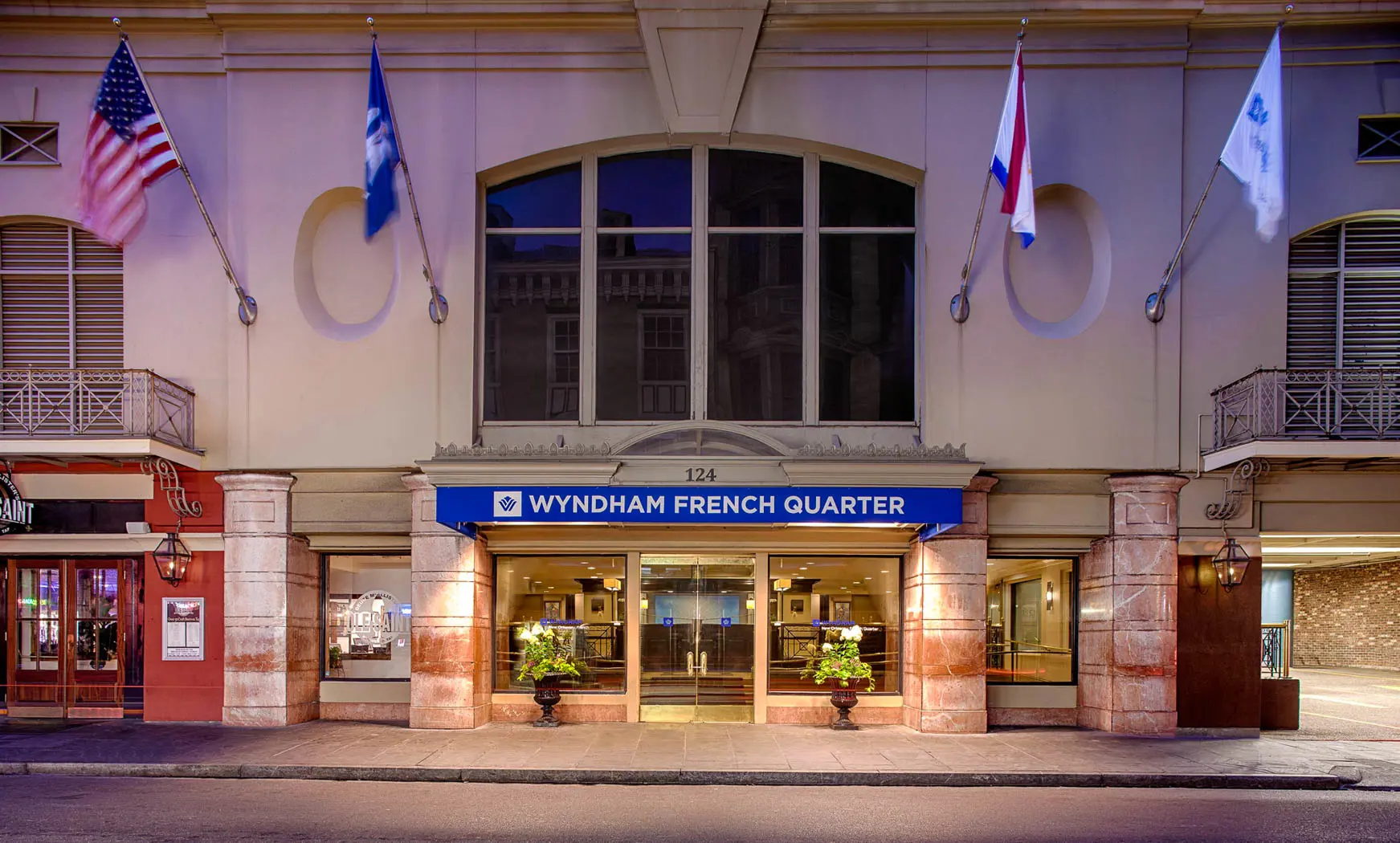 Entrance to the Wyndham French Quarter hotel, featuring a well-lit façade with stone pillars, a blue sign above the glass doors, and several flags flying above
