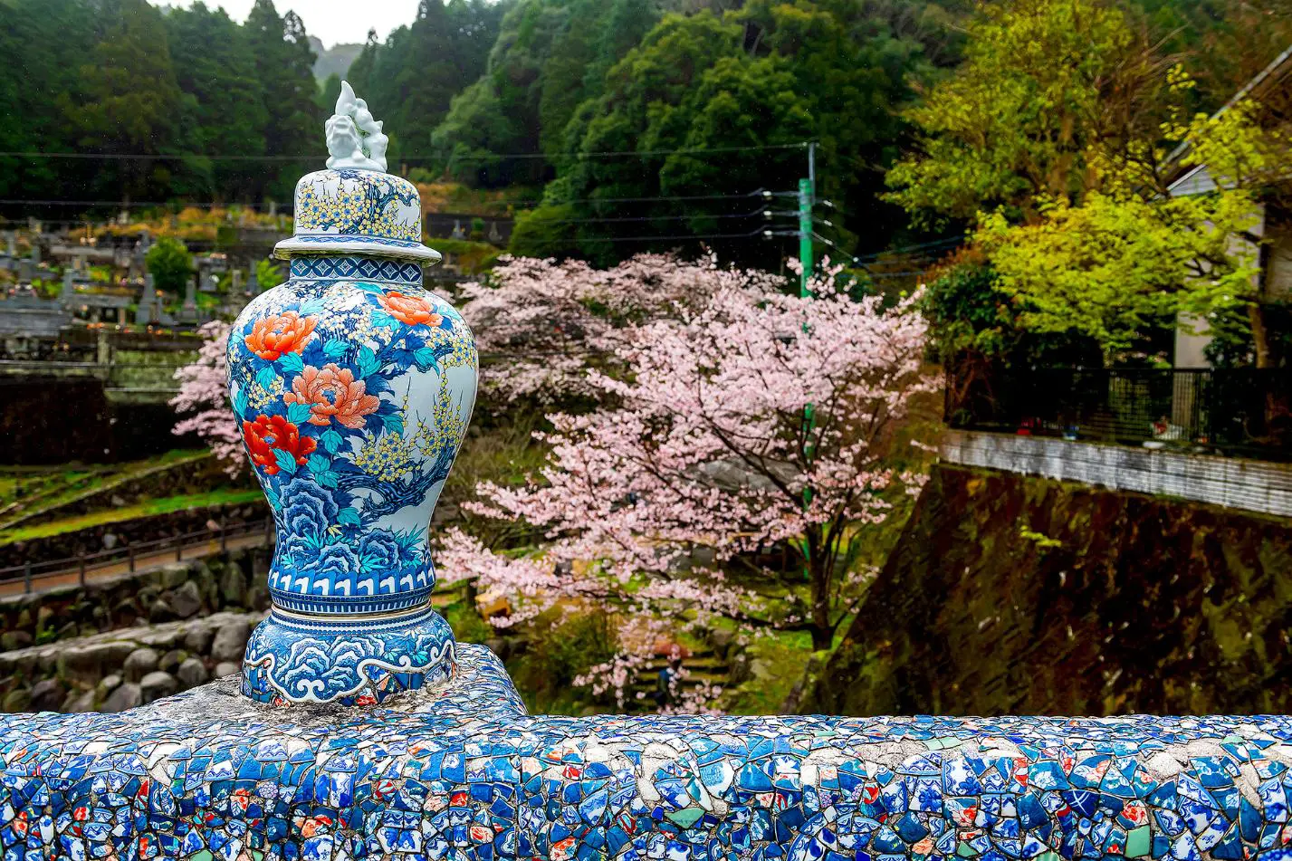 A decorative porcelain vase on a mosaic-tiled bridge in Okawachiyama village, with cherry blossoms and forested hills in the background