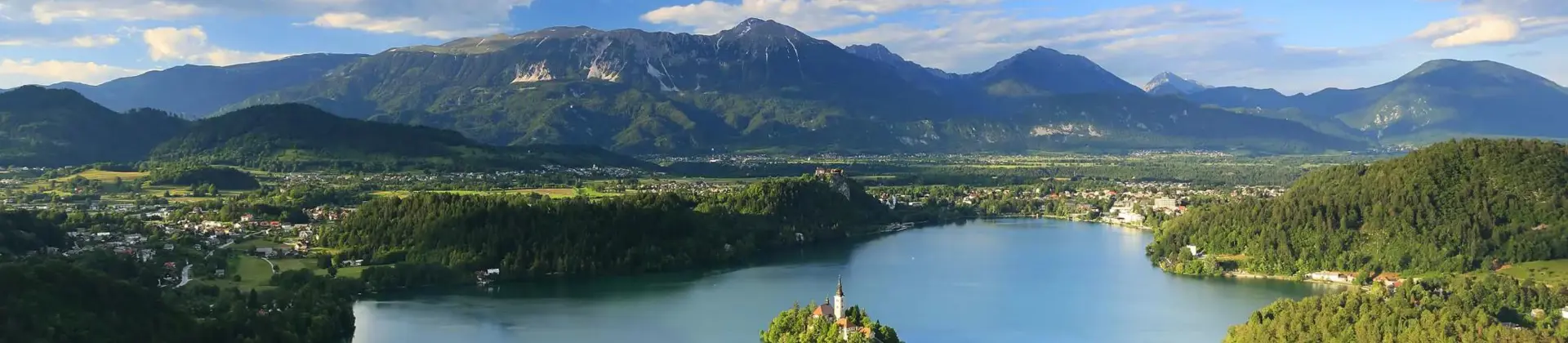 Wide angle shot of Lake Bled, showing its forested surrounding areas, and mountains in the distance. In the middle of the lake, there is a small island with trees and a towered building