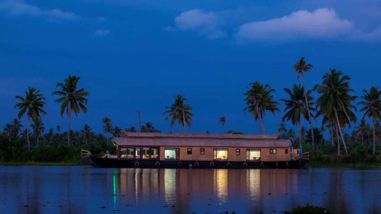 A traditional wooden houseboat floats on calm waters surrounded by lush green palm trees under the night sky in Kerala, India