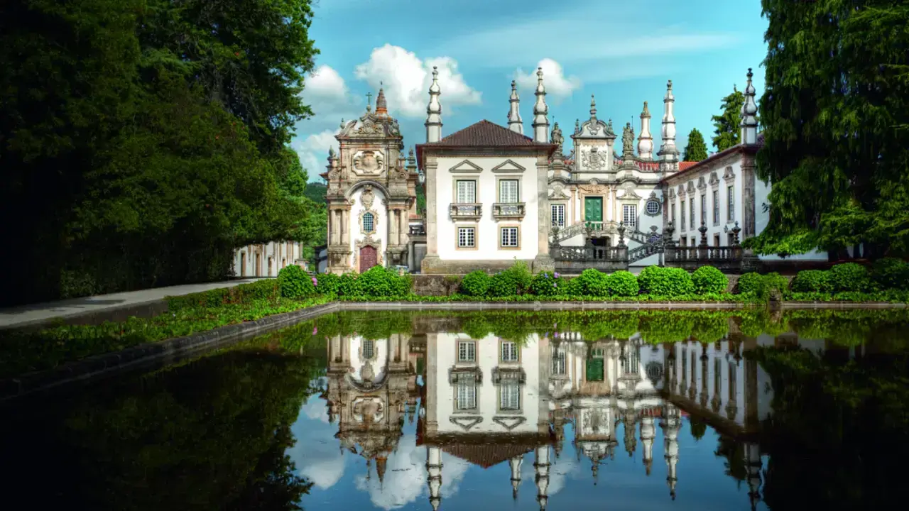 Front view of the ornate Mateus Palace in Portugal, with its baroque architecture reflected in a still pond, surrounded by lush greenery under a bright blue sky