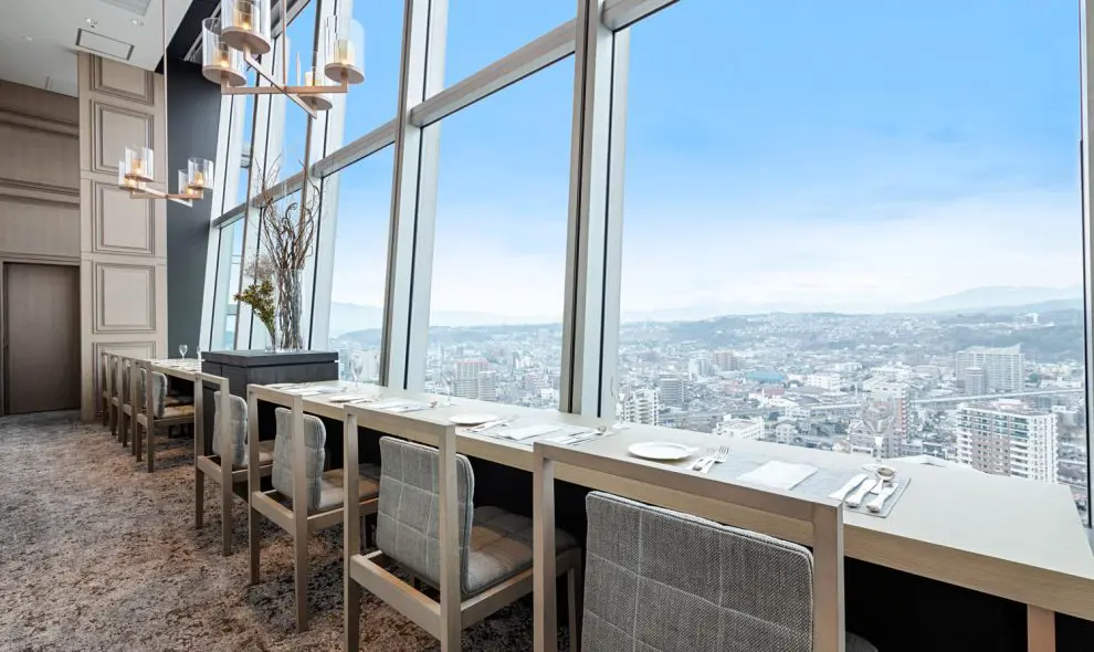 Elegant dining area at Hotel Nikko Ōita Oasis Tower, with floor-to-ceiling windows offering panoramic views over Ōita city, set with modern chairs and white table settings