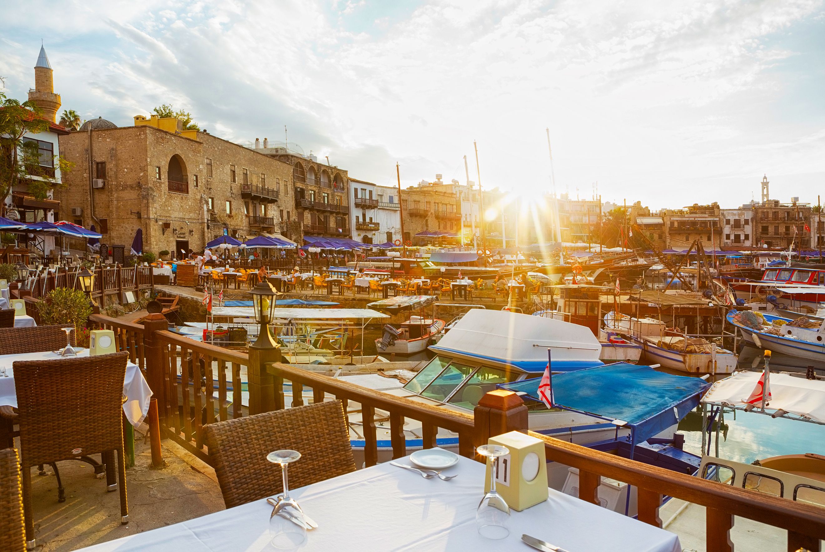 View of a marina from a restaurant, with a laid table in the forefront behind a wooden barrier dividing the boats on the water and the land. Other buildings and restaurants can be seen on the other side, with blue umbrellas and outdoor seating. The sun is poking over the buildings, with beams shining into the camera