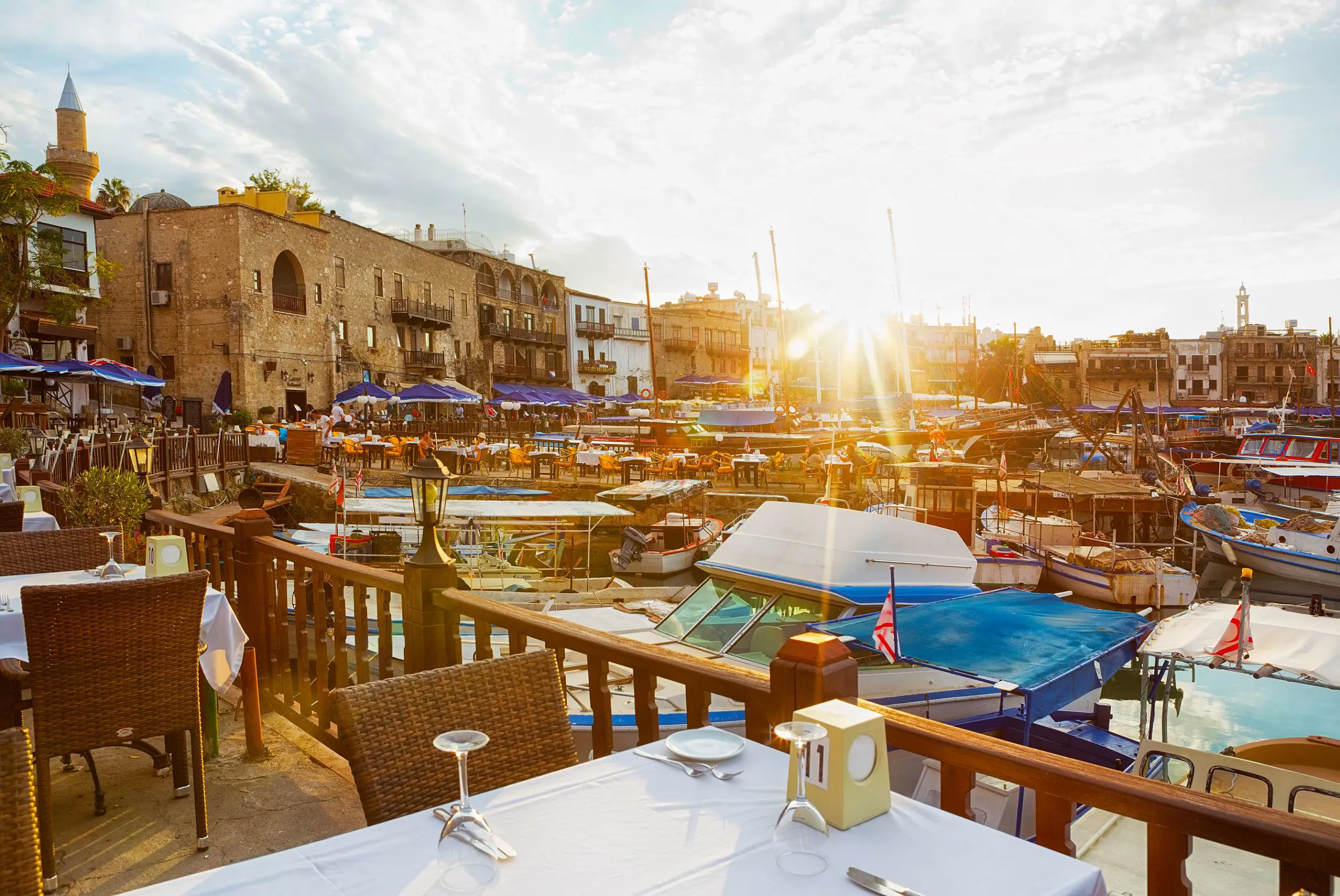 View of a marina from a restaurant, with a laid table in the forefront behind a wooden barrier dividing the boats on the water and the land. Other buildings and restaurants can be seen on the other side, with blue umbrellas and outdoor seating. The sun is poking over the buildings, with beams shining into the camera