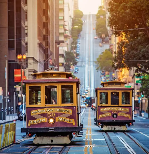 Two traditional San Francisco cable cars in yellow and maroon colours descending a steep hill with parked cars either side of the street
