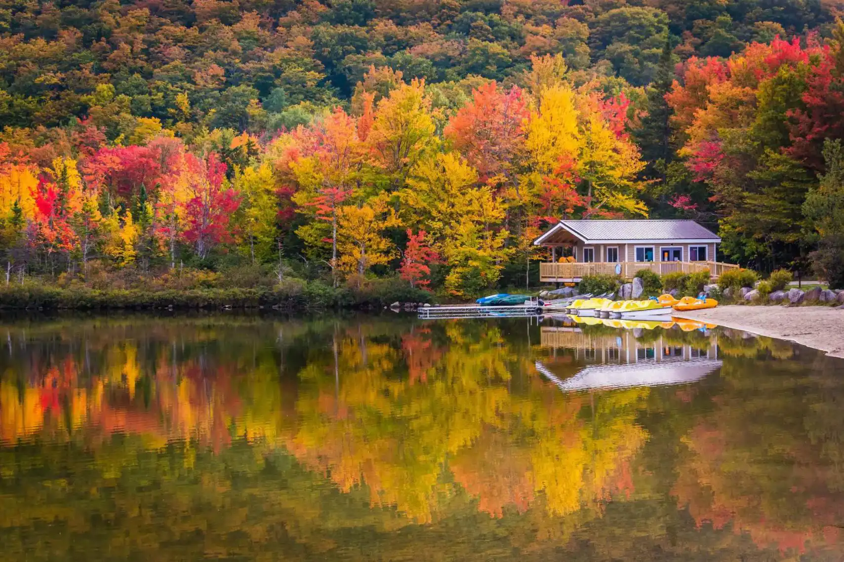 A peaceful lake reflects vibrant autumn trees and a cosy wooden cabin with kayaks on the shore in New Hampshire, USA