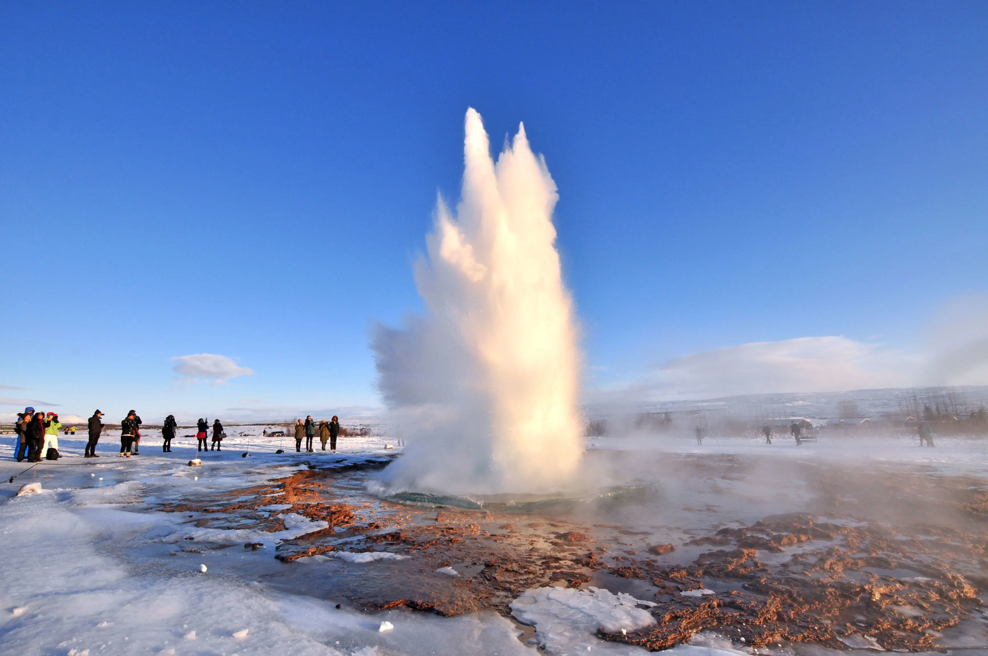 Erupting geysur, surrounded by rocks and ice. People stood watching