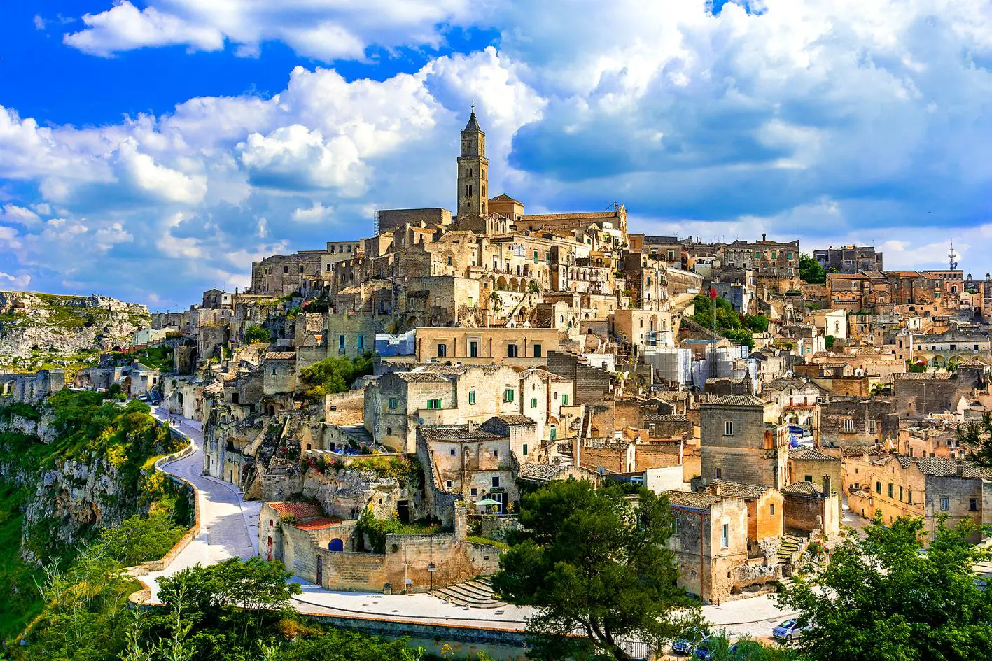 A panoramic view of the historic stone town of Matera, Italy, with ancient buildings stacked on a hillside under a partly cloudy blue sky