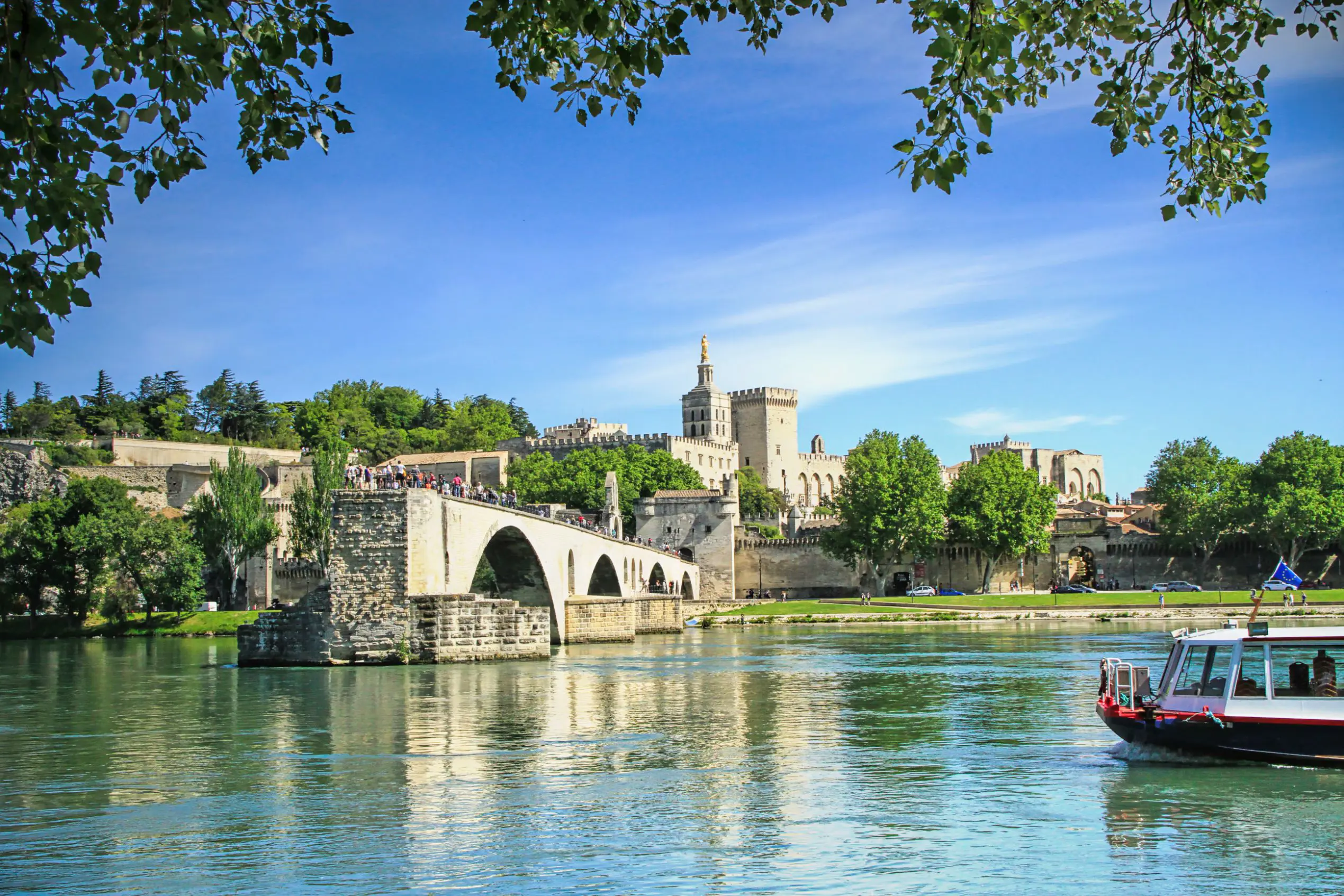 View of the Pont D’Avignon bridge, with the river water in the forefront and a boat coming from the right. In the distance, on the other side of the bridge is a grey palace. The sky is blue.