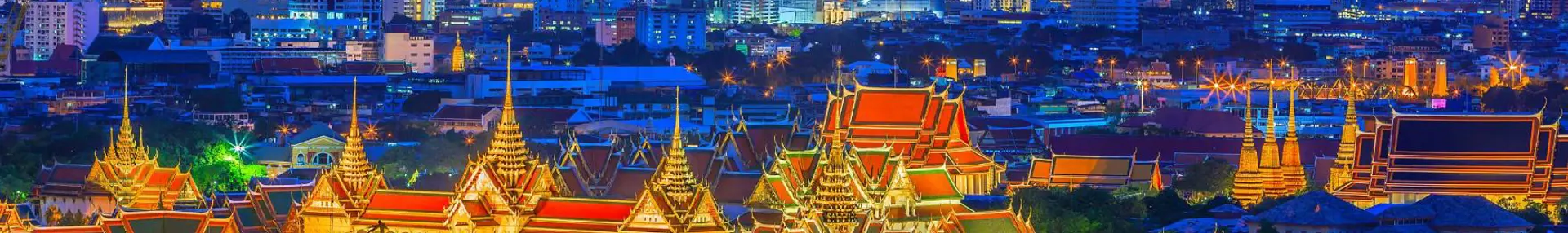 Distant view of the Grand Palace in Bangkok lit up at night, with its golden spires and ornate rooftops surrounded by softly illuminated temples and skyscrapers