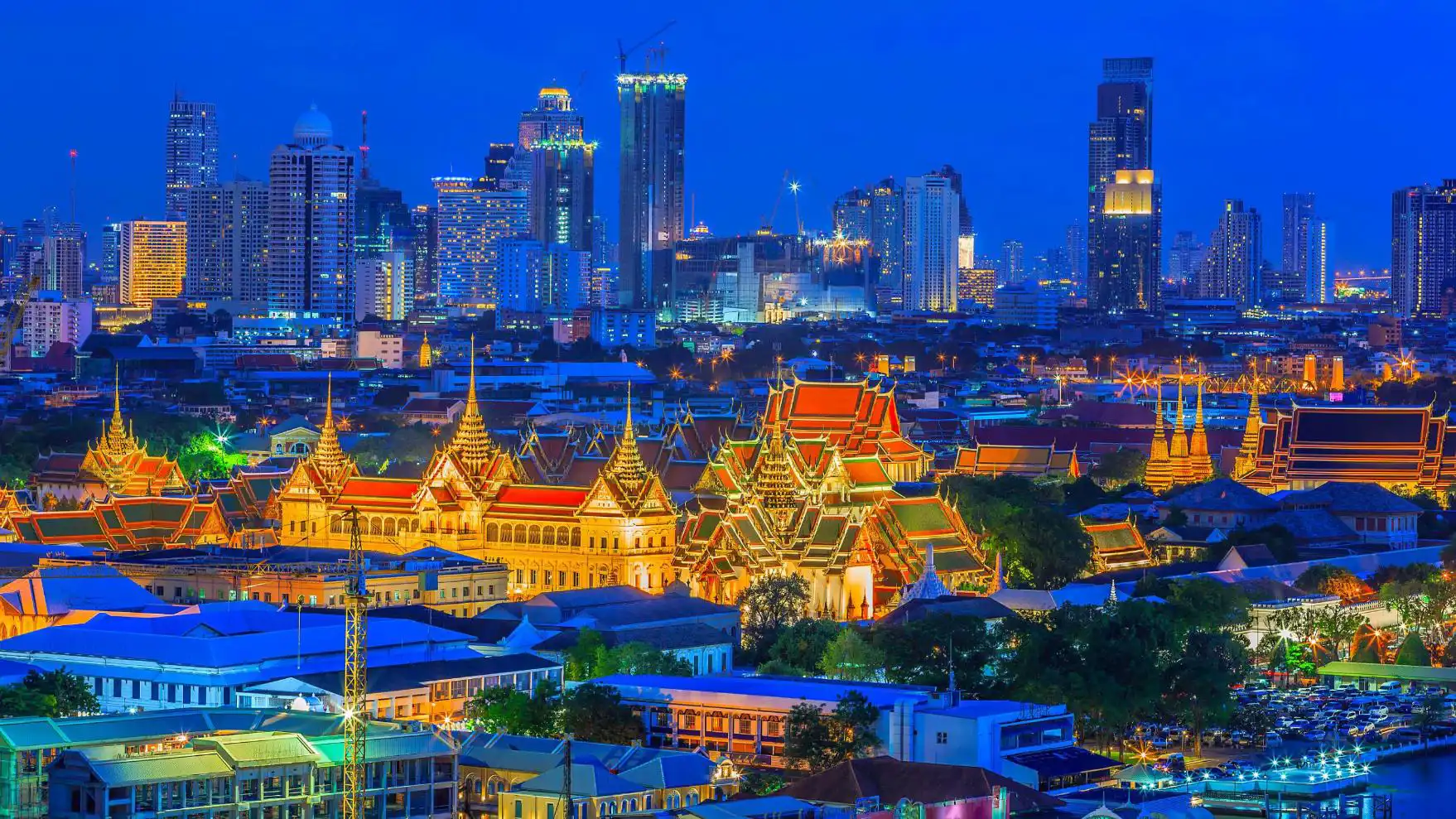 Distant view of the Grand Palace in Bangkok lit up at night, with its golden spires and ornate rooftops surrounded by softly illuminated temples and skyscrapers