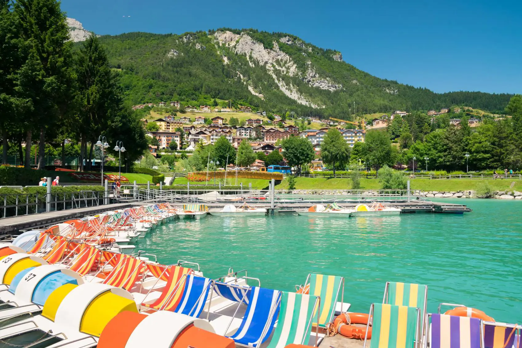 Colourful pedal boats line a dock on Lake Molveno in Italy with a village and forested mountains in the background