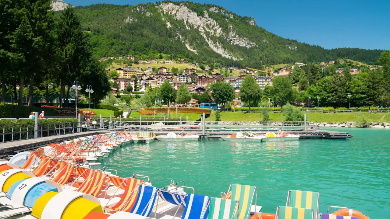 Colourful pedal boats line a dock on Lake Molveno in Italy with a village and forested mountains in the background