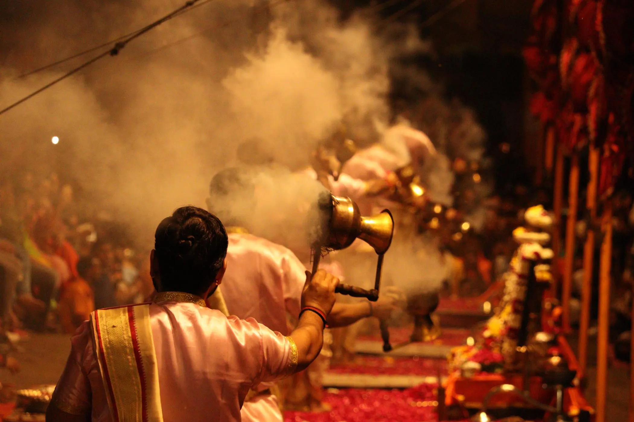 A close view of priests performing the Ganga Aarti ritual in Varanasi, holding brass lamps with incense smoke swirling in the air amid a reverent evening crowd