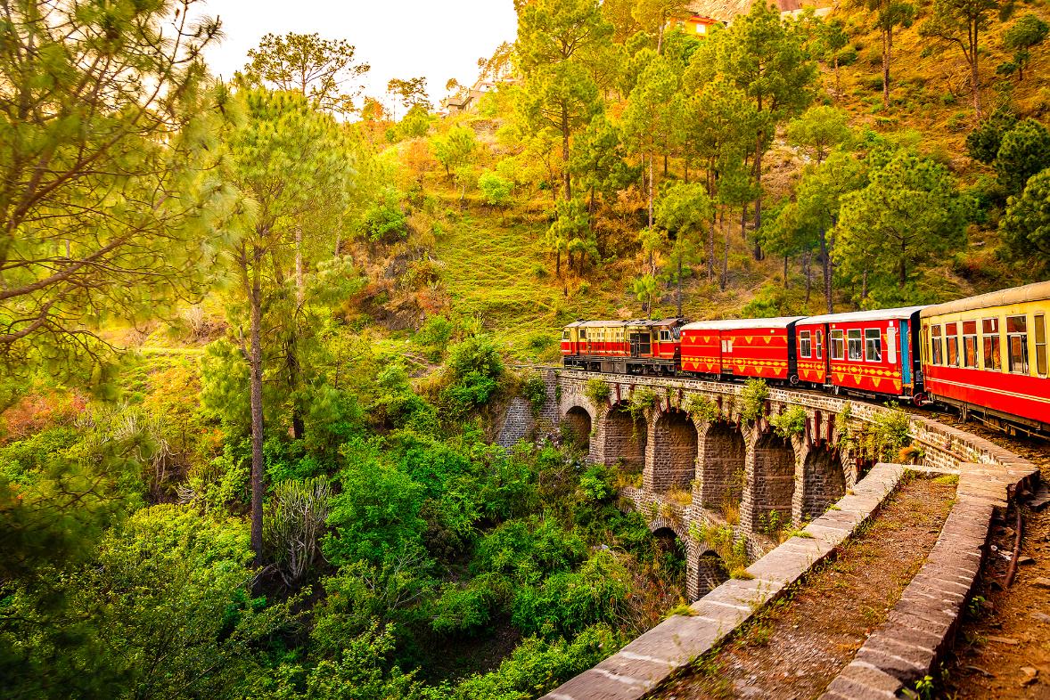 The heritage toy train crossing a railway bridge on the Kalka–Shimla route, surrounded by lush green hills and trees