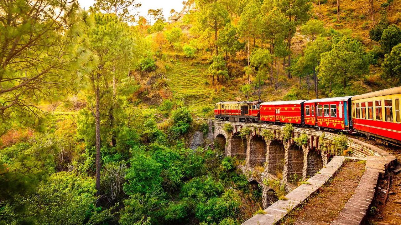 The heritage toy train crossing a railway bridge on the Kalka–Shimla route, surrounded by lush green hills and trees