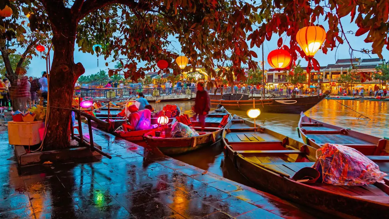 Traditional wooden boats moored on a tranquil river in Hoi An, Vietnam, with colourful lanterns hanging from tree branches overhead