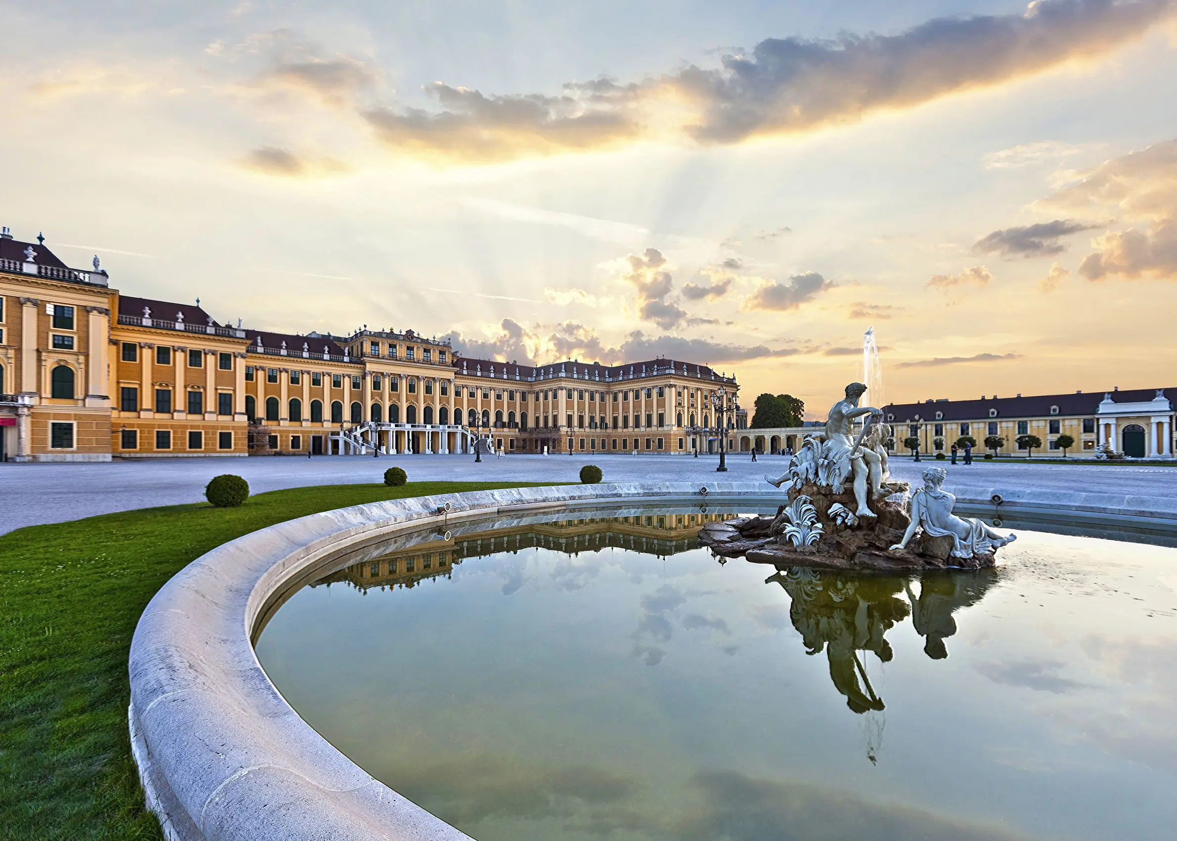 A pond with a fountain with a white statue of people sitting on a rock in the right forefront, behind is Schoenbrunn Palace, a yellow and orange, wide building in front of a bright sky with a yellow hue.