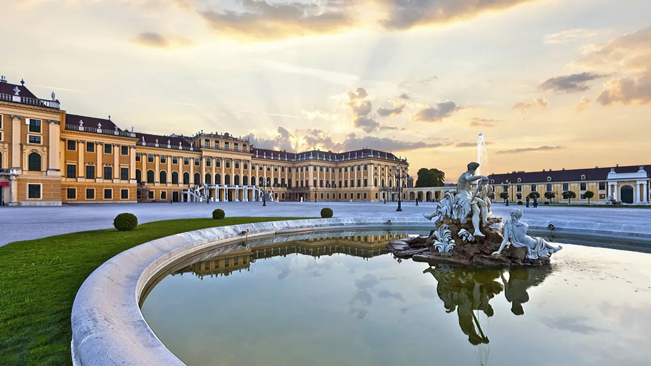 A pond with a fountain with a white statue of people sitting on a rock in the right forefront, behind is Schönbrunn Palace, a yellow and orange, wide building in front of a bright sky with a yellow hue.